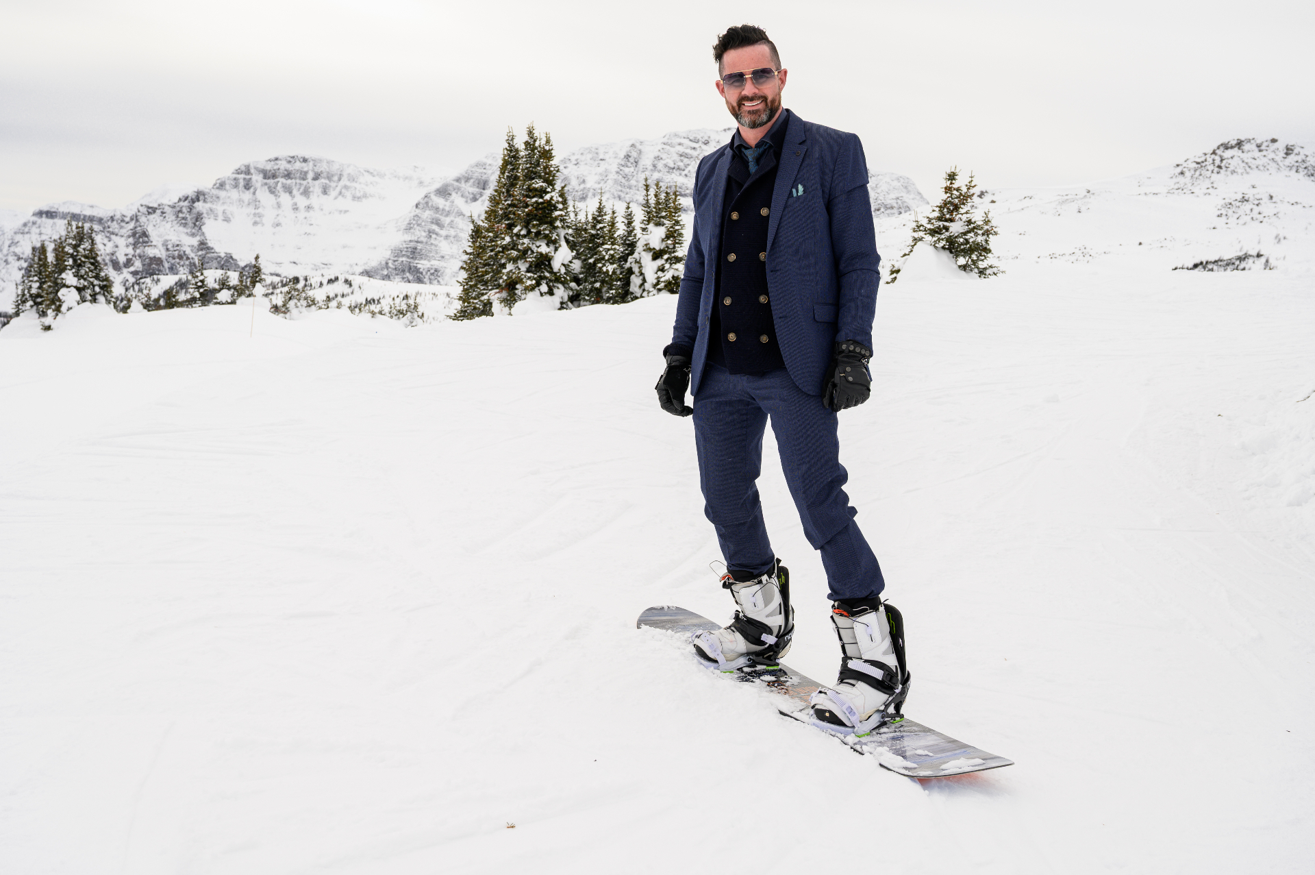 Paul in a navy suit on a snowboard, on a groomed slope with mountains behind