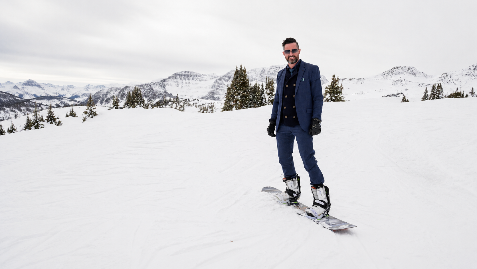 Paul in a navy suit on a snowboard, on a groomed slope with mountains behind